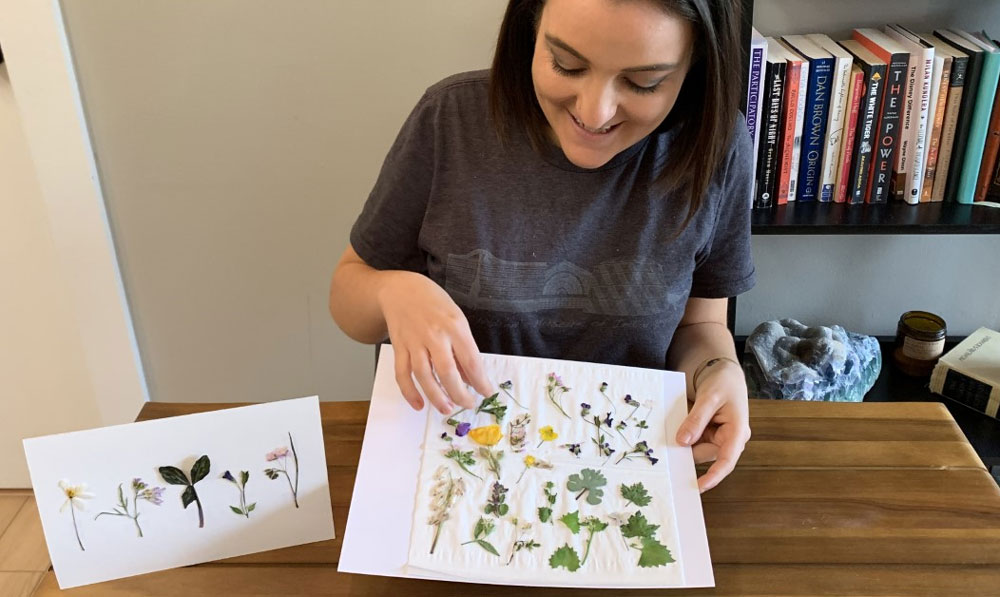 Public and Youth Programs Coordinator Lindsey DeLorey displaying a graphic of wildflowers with an example of framed pressed wildflowers beside her.